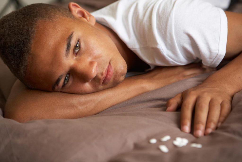 lagunashoresrecovery How Normal People End Up Addicted to Opioids photo of Depressed Teenage Boy Lying In Bedroom With Pills