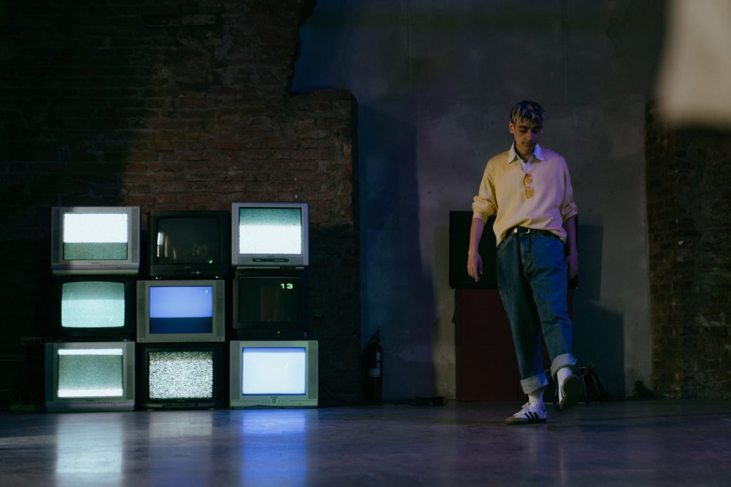 man in yellow polo shirt and blue jeans standing near black old tv screens